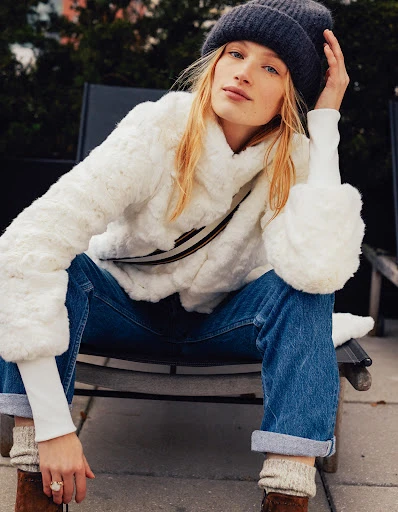 Blonde model poses in a white faux-fur jacket, jeans, and a gray beanie, seated outdoors on a black chair, in a medium portrait shot.