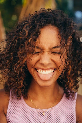 A portrait of a young adult woman with curly brown hair sticking her tongue out and squinting her eyes while wearing a pink shirt.