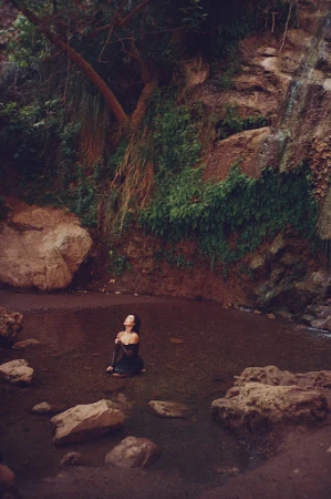 Woman in a black dress kneeling in a shallow pool of water near a rocky waterfall, photographed by Kayla Varley