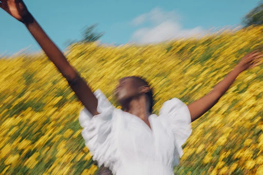 An adult woman in a white dress stands in a yellow flower field with her arms raised against a blue sky in a blurry, dreamy, motion-blurred style.