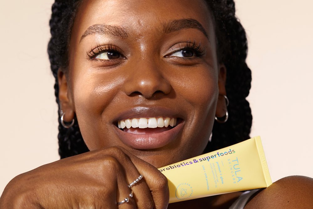 Close-up of a smiling African American woman with yellow skincare tube, silver hoop earrings and rings against a cream background.