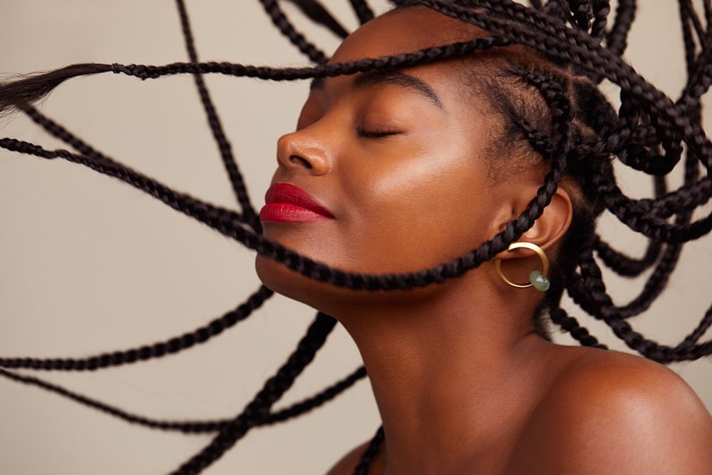 Close-up of a young Black woman with eyes closed, glossy red lips, and box braids, set against a plain background.