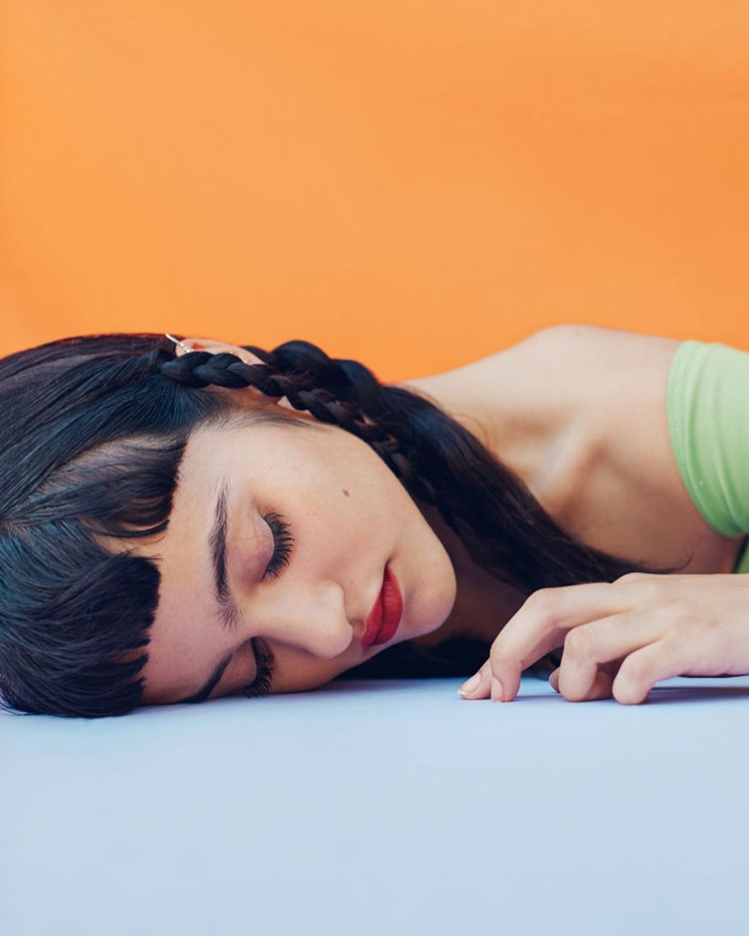 Close-up studio shot of a young woman with dark hair and red lipstick lying down, with an orange background and pale blue surface.