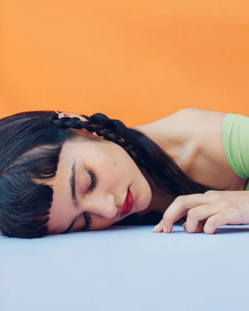 Close-up studio shot of a young woman with dark hair and red lipstick lying down, with an orange background and pale blue surface.