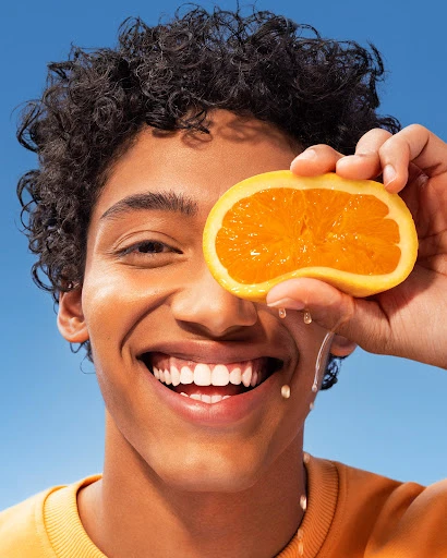 Smiling young adult man holds a half-cut orange slice to his eye with water droplets against a clear blue sky in a close-up shot.