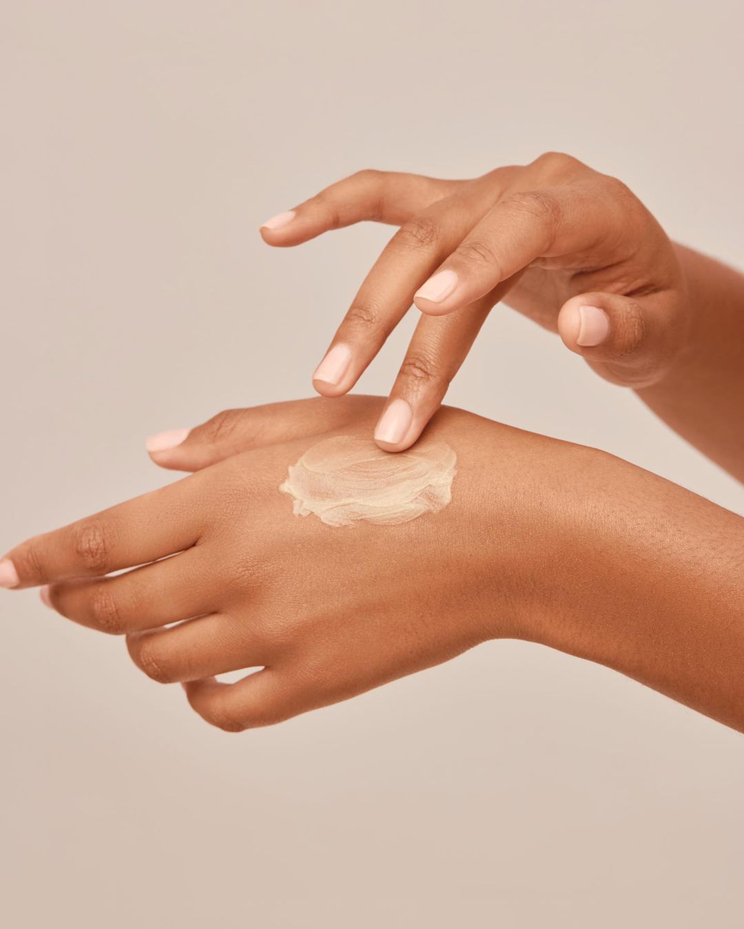 Close-up of dark-skinned hands applying cream, emphasizing skincare, moisturizing, and natural beauty against a neutral backdrop.