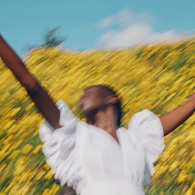 Woman in white dress with arms raised stands in field of yellow flowers under blue sky, captured with motion blur effect.