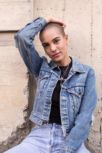 Portrait of a shaved woman in a blue denim jacket with her hand raised against a textured stone wall