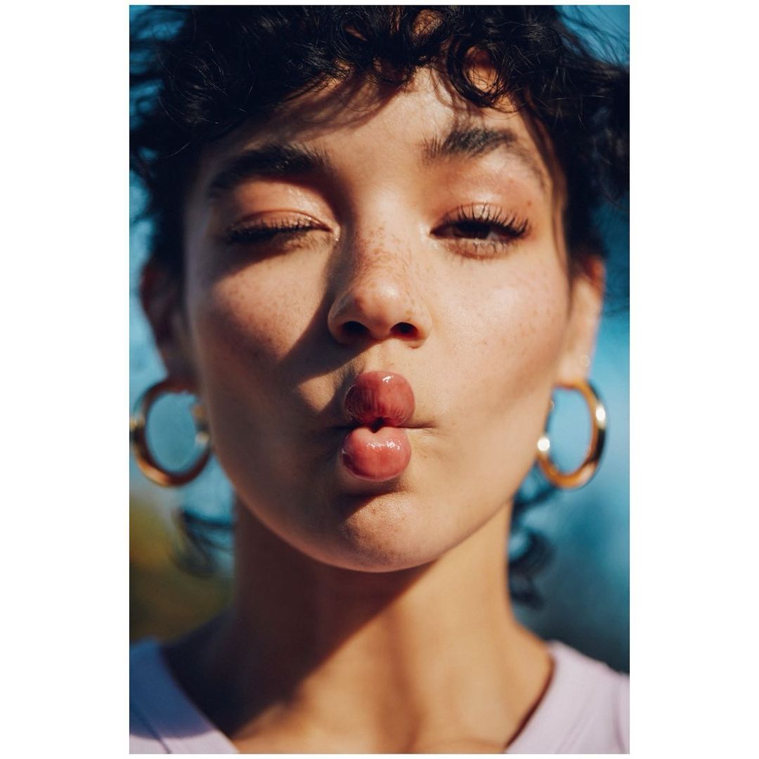 Close-up portrait of a young woman with freckles and dark curly hair puckering her lips, accessorized with gold hoop earrings against a blue backdrop.