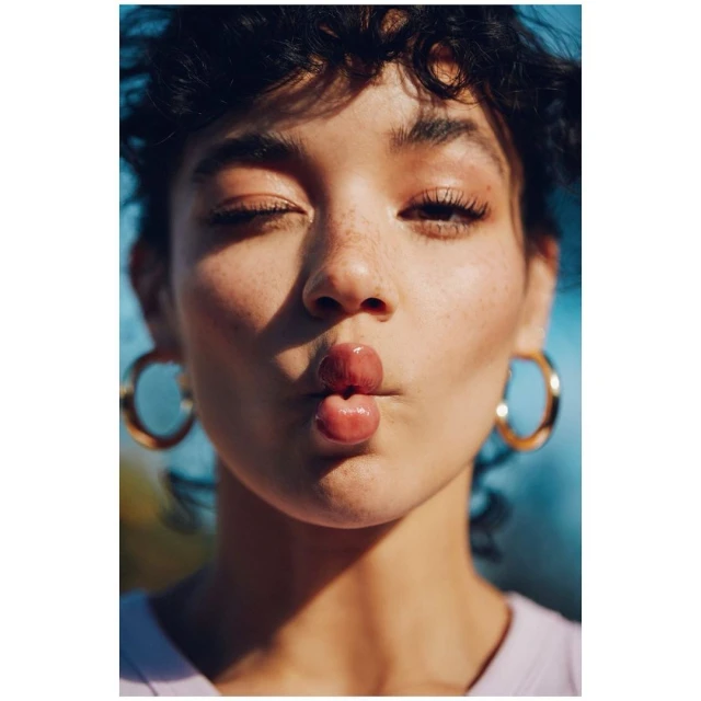 Close-up portrait of a young woman with freckles and dark curly hair puckering her lips, accessorized with gold hoop earrings against a blue backdrop.