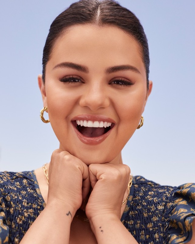 Selena Gomez smiling broadly with hands under chin, wearing gold hoop earrings and patterned top against clear blue sky background