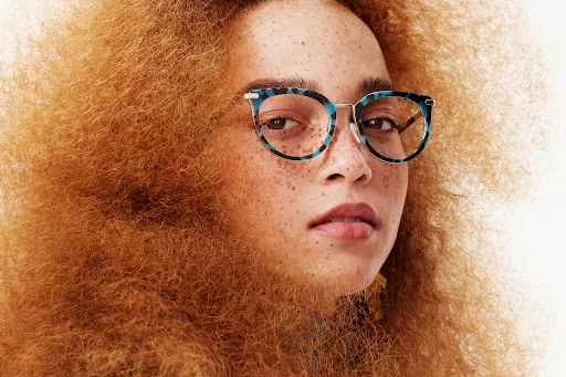 Close-up portrait of a woman with freckles and voluminous curly ginger hair wearing blue tortoiseshell eyeglasses