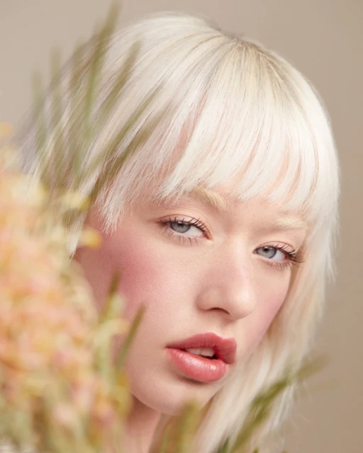 Close-up portrait of a woman with platinum blonde hair and pale skin with a peach-toned floral element in the foreground.