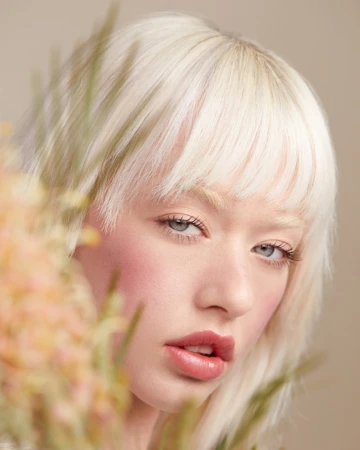Close-up portrait of a woman with platinum blonde hair and pale skin with a peach-toned floral element in the foreground.