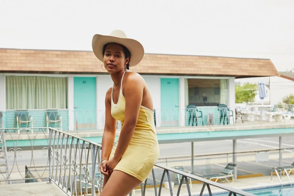 Woman in yellow dress and cowboy hat sitting on balcony railing in front of vintage motel with turquoise doors and a swimming pool.