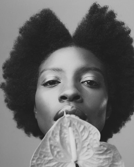 Black and white portrait of a Black woman with an afro holding a white anthurium flower to her lips, close-up with a neutral background.