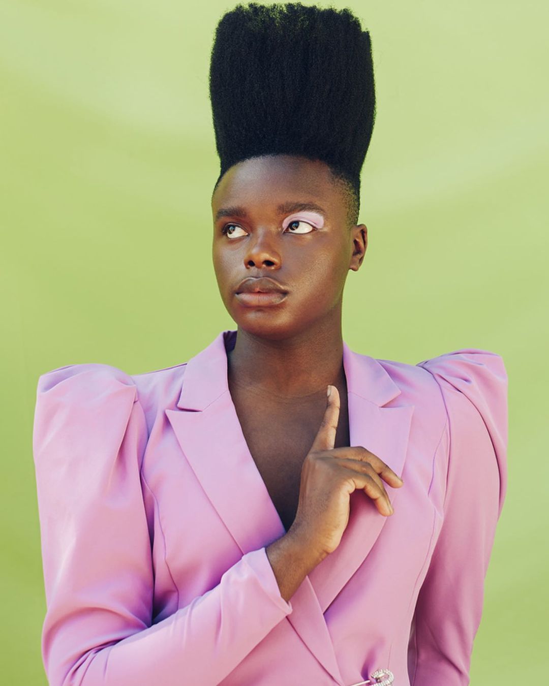 Portrait of a Black model with a high-top fade, wearing a pink blazer in front of a lime green background, looking to the side.