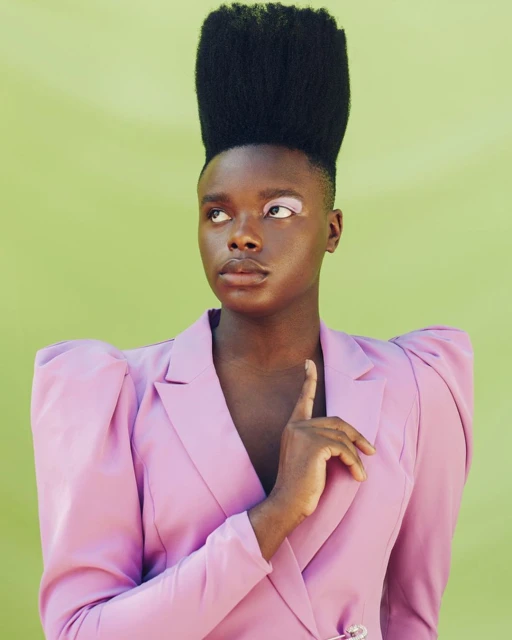 Portrait of a Black model with a high-top fade, wearing a pink blazer in front of a lime green background, looking to the side.