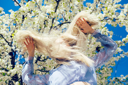 A person with blonde hair obscures their face with their hands while standing in front of a blooming tree and a clear blue sky outdoors.