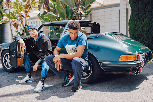 Two men sit on and next to a vintage turquoise Porsche car on a city street, both wearing Puma branded matching apparel and shoes.