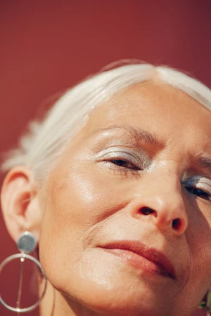 Close-up portrait of an elderly woman with silver hair wearing large hoop earrings against a dark red background.