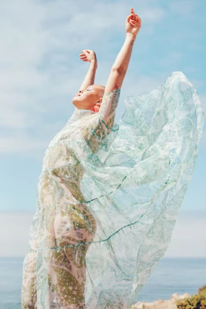 Woman in a sheer patterned green and white dress with arms raised outdoors against an ocean background