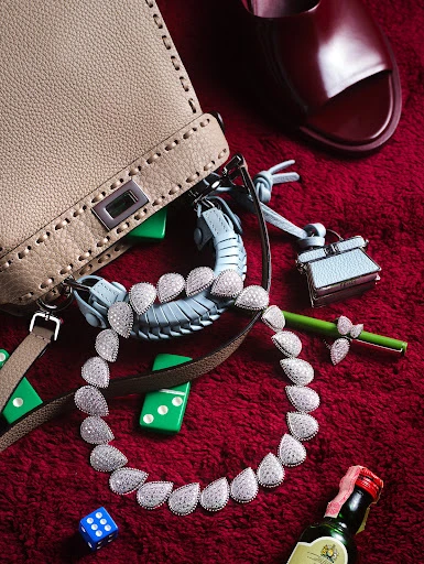 Fashion still life featuring a beige purse, jewelry necklace, red mules, dominoes, and a miniature liquor bottle on a red surface.