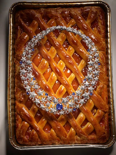 Overhead studio shot of a lattice-crust apricot pie in a metal baking pan with a star-shaped silver and blue gemstone necklace on top.