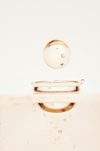Close-up studio shot of a clear liquid sphere suspended above a droplet on a surface, featuring soft, neutral tones and subtle light play.