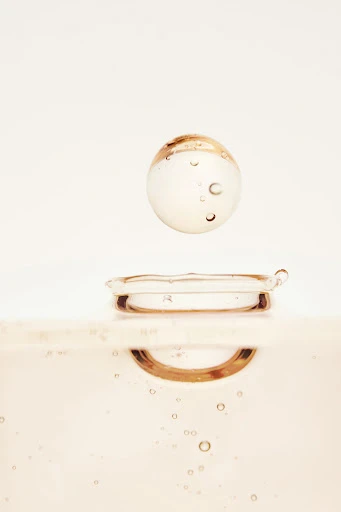Close-up studio shot of a clear liquid sphere suspended above a droplet on a surface, featuring soft, neutral tones and subtle light play.