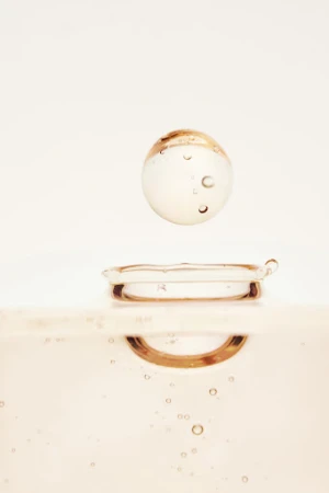 Close-up studio shot of a clear liquid sphere suspended above a droplet on a surface, featuring soft, neutral tones and subtle light play.