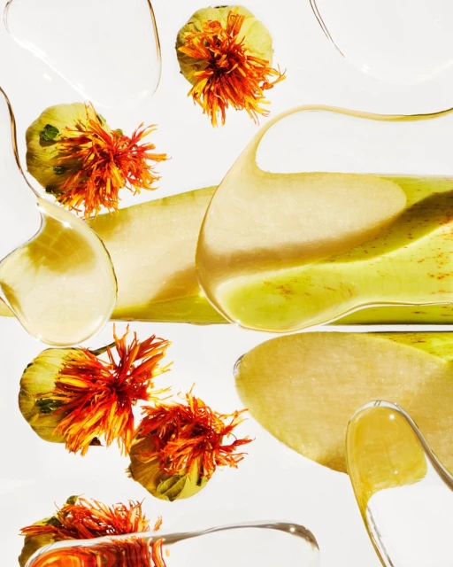 Abstract still life featuring safflower blooms, green seed pods, and translucent gel drops against a clean, minimalist white backdrop.