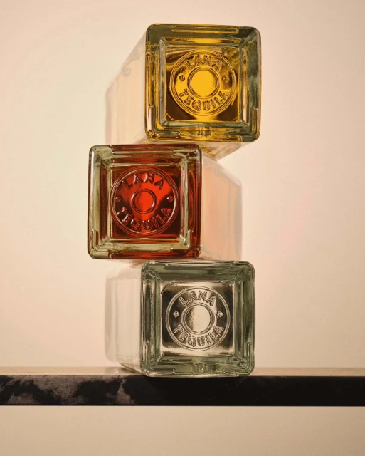 Three square tequila bottles stacked vertically, displaying amber, reddish, and clear liquids, casting shadows against a cream backdrop.