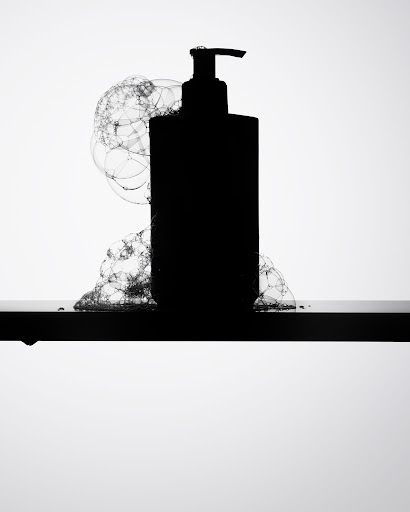 Silhouette of a pump bottle sits on a black surface, surrounded by soap bubbles against a bright white background in a studio shot.