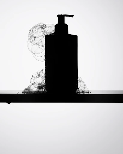 Silhouette of a pump bottle sits on a black surface, surrounded by soap bubbles against a bright white background in a studio shot.