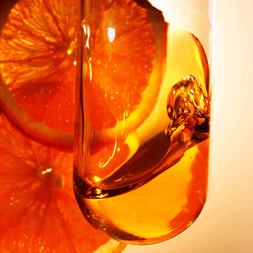 Close-up studio shot of a clear glass filled with an orange-colored liquid, backlit with slices of oranges in the background.