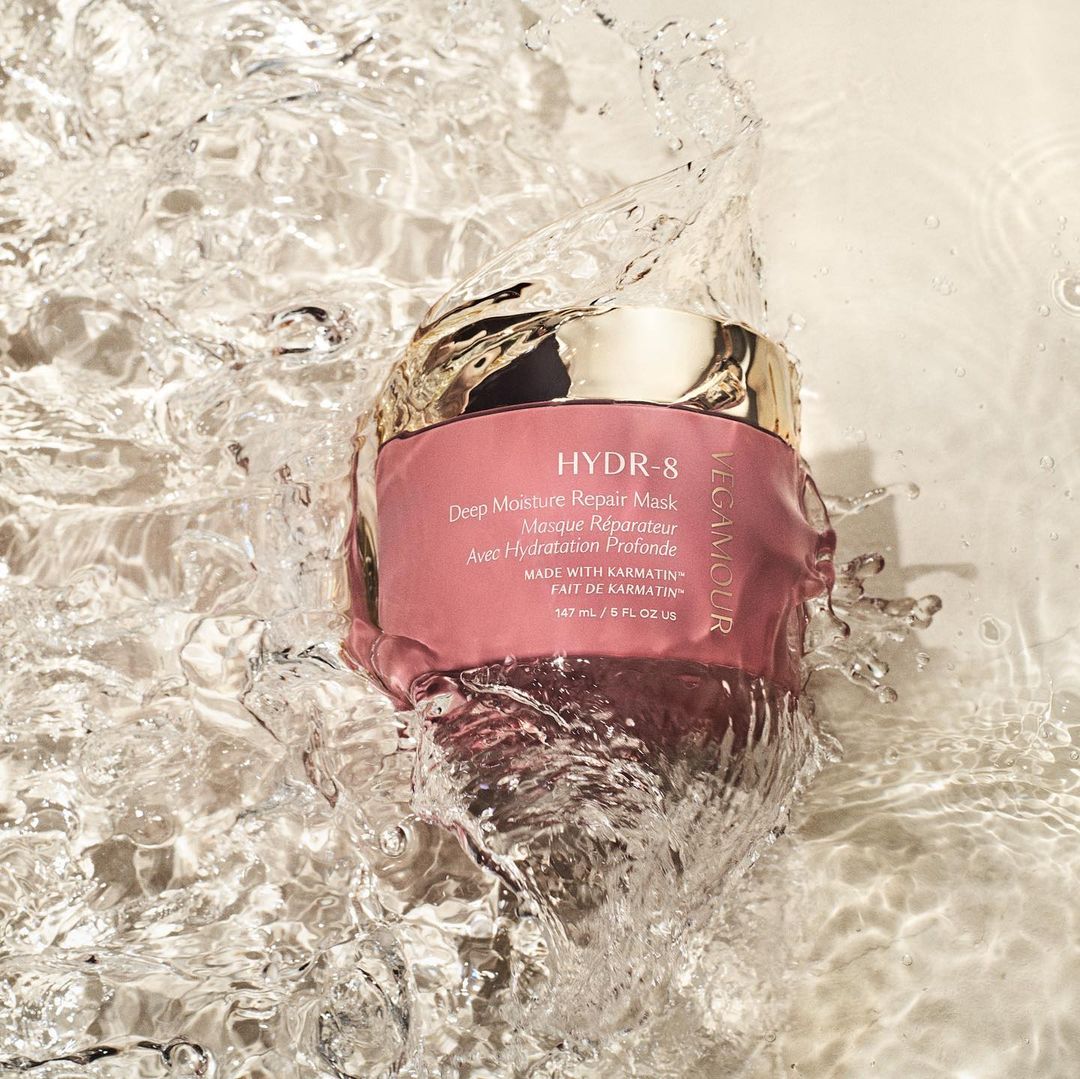Vegamour HYDR-8 Deep Moisture Repair Mask is showcased in water with splashes, featuring a pink container and gold lid against a bright background.