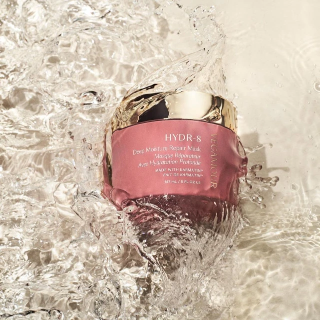 Vegamour HYDR-8 Deep Moisture Repair Mask is showcased in water with splashes, featuring a pink container and gold lid against a bright background.