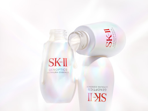 Three SK-II GenOptics Ultraura Essence bottles stacked, one lying on top with product dripping, white background, product shot.