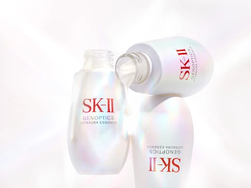 Three SK-II GenOptics Ultraura Essence bottles stacked, one lying on top with product dripping, white background, product shot.