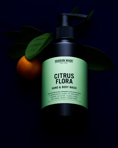 Still life shot of black bottle of Hudson Made Citrus Flora hand and body wash next to an orange and green leaves against a dark backdrop.