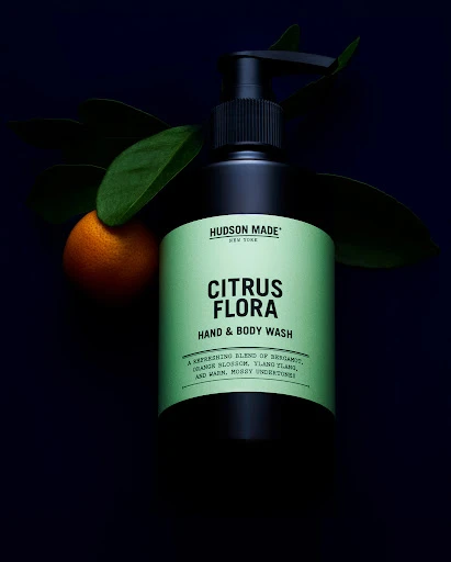 Still life shot of black bottle of Hudson Made Citrus Flora hand and body wash next to an orange and green leaves against a dark backdrop.