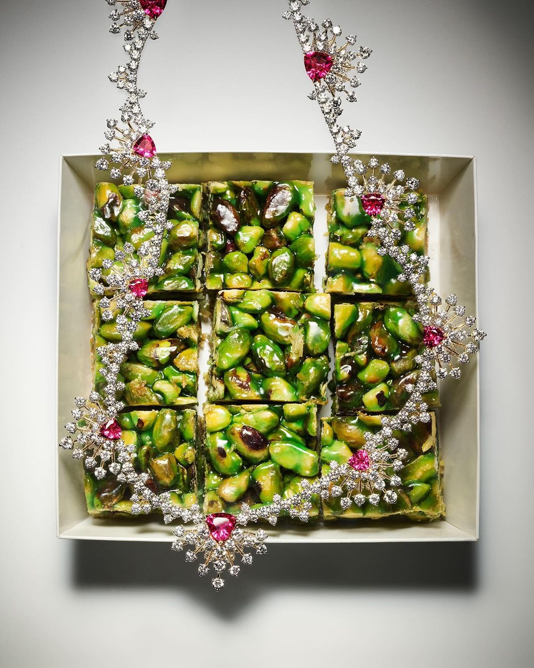 Overhead shot of pistachio squares arranged in a square baking pan and adorned with a decorative diamond and ruby necklace.