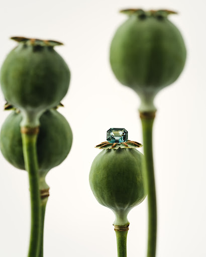 Still life shot of a blue gemstone atop a green poppy seed pod against a white background, with other pods softly blurred behind it.