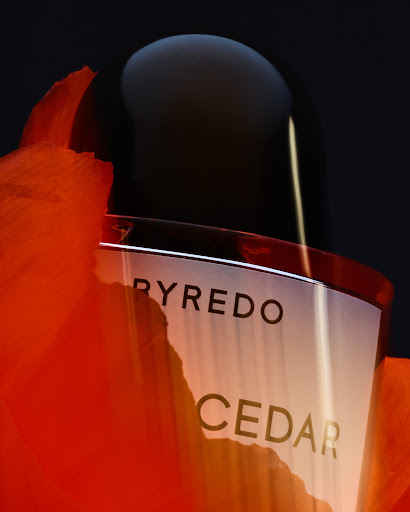 Close-up of a Byredo Cedar perfume bottle with orange petals against a black background, showcasing its sleek design and packaging.