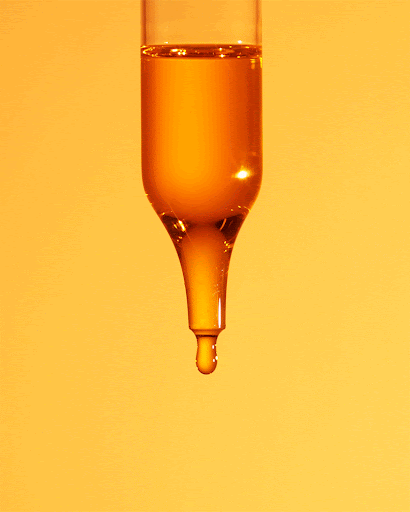 Close-up of a transparent glass dropper filled with golden yellow liquid with a single drop forming at the tip, on a yellow background.