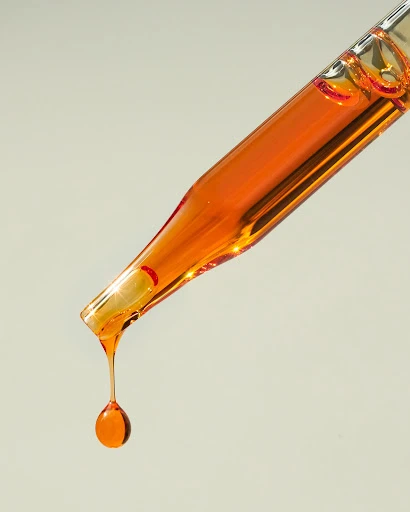 Close-up studio shot of an orange liquid dripping from a clear glass pipette against a soft grey backdrop, minimal and clean.