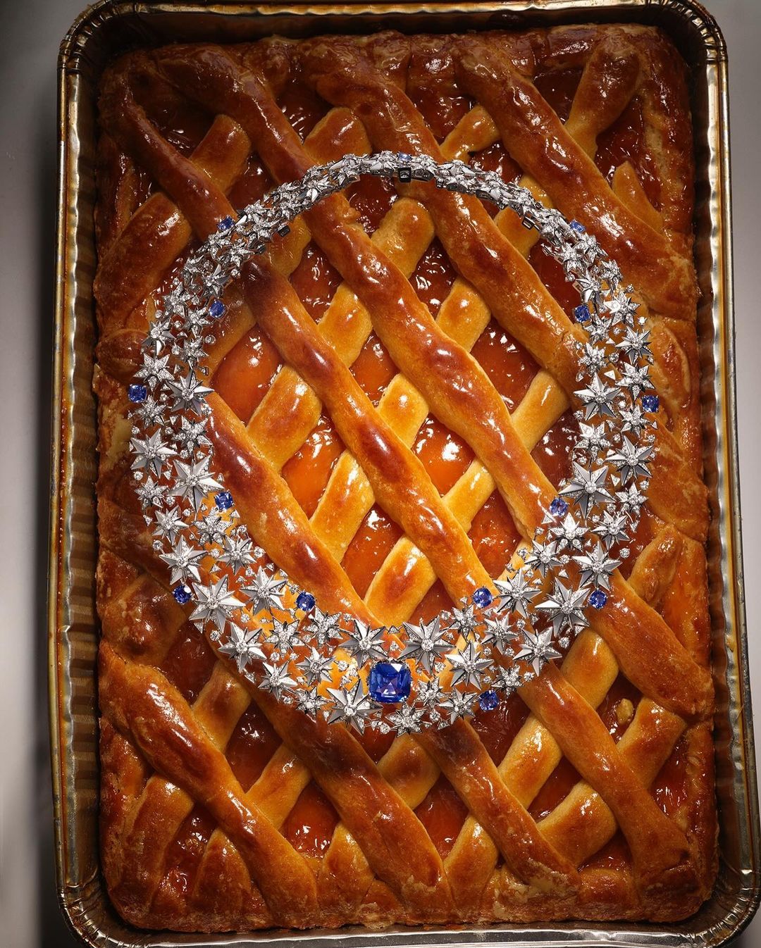 Still life photograph of a diamond and sapphire necklace displayed on a lattice-crust apricot tart in a baking pan, studio lighting.