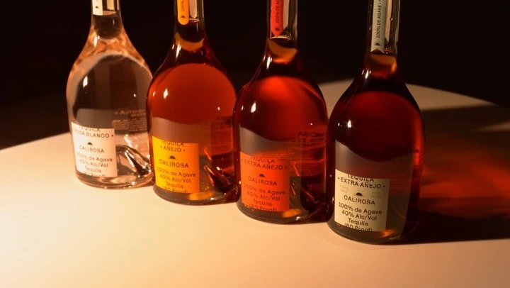 Four bottles of Calirosa tequila varieties (Rosa Blanco, Anejo, Extra Anejo) stand in a row under warm spotlighting in a studio setting.