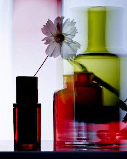 Still life shot of a single white daisy in a black vase, green glass and red glass bottles behind, against a white background.